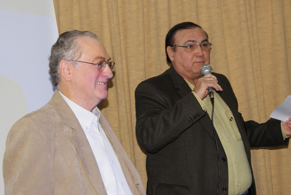 Dois homens de pé lado a lado diante de uma cortina clara; o da direita, de terno escuro e camisa verde, fala ao microfone segurando uma folha, e o da esquerda, de paletó bege, olha para ele.