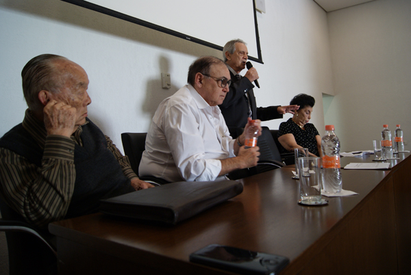 Mesa da Assembleia Geral Ordinária: quatro pessoas sentadas a uma bancada de madeira com garrafas de água e copos; ao fundo, um homem de pé fala ao microfone diante da tela de projeção.