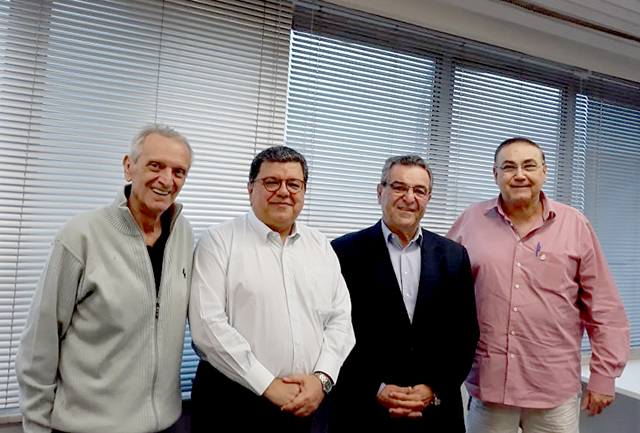Quatro homens em pé, lado a lado, sorriem para a foto numa sala de reuniões com persianas ao fundo.