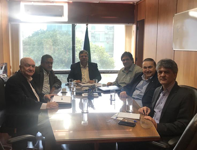 Seis homens sentados em torno de uma mesa de reunião com tampo escuro, em sala com painéis de madeira e uma bandeira do Brasil junto à janela; sobre a mesa, copos, pastas e documentos.