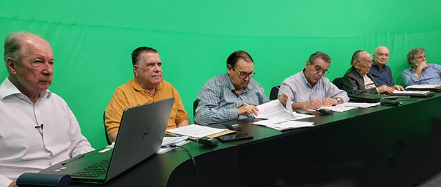 Sete dirigentes sentados em fila à mesa do estúdio, com notebooks e papéis à frente, diante do fundo verde.