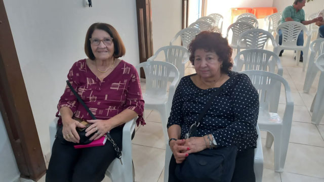 Duas associadas sentadas em cadeiras brancas sorriem para a foto no salão onde ocorreu a reunião.