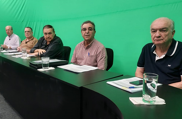 Cinco dirigentes sentados lado a lado à mesa da assembleia, com papéis e copos d’água à frente, diante do fundo verde do estúdio.