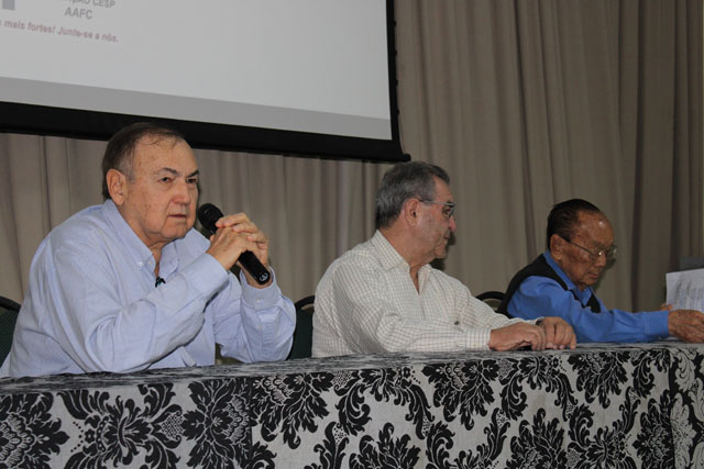 Três dirigentes à mesa de abertura, coberta por toalha estampada; o primeiro fala ao microfone e os outros dois acompanham.