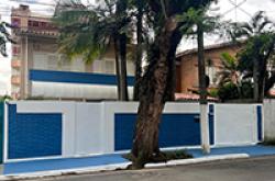 Fachada da sede da regional: muro branco com faixa azul e portões azuis, uma palmeira e uma árvore de tronco grosso na calçada.