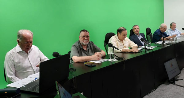 Seis dirigentes sentados à mesa do estúdio durante a assembleia virtual, com microfones e notebooks à frente, diante do fundo verde.
