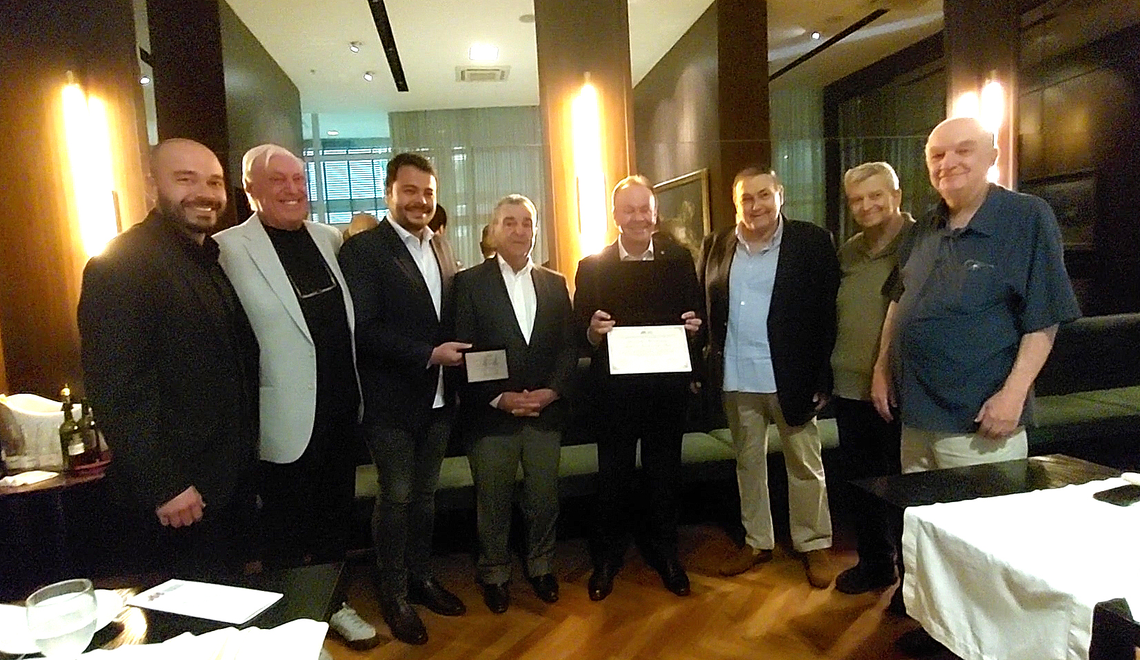 Nove homens em pé, lado a lado, num salão de restaurante; ao centro, dois deles seguram a placa e o diploma entregues na homenagem.