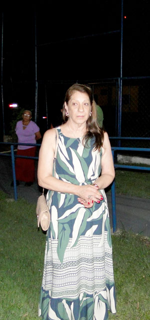 Mulher de vestido longo estampado em verde e branco, em pé na área externa da pousada à noite; ao fundo, outra convidada junto ao alambrado.