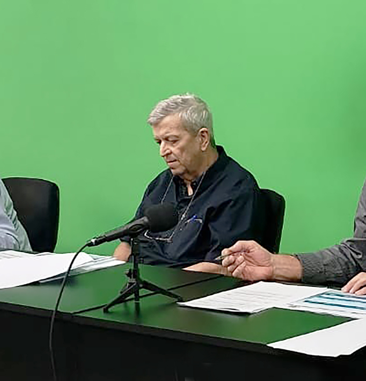 Homem de cabelos brancos e camisa escura lê documentos diante de um microfone, sentado à mesa do estúdio com painel verde ao fundo; ao lado, as mãos de outro participante sobre as folhas.